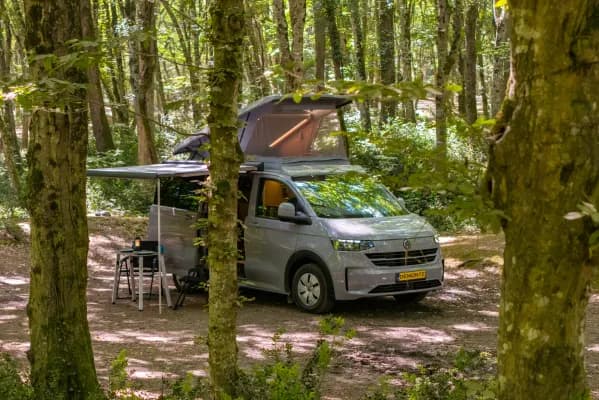 Demonte-Camper zwischen den Bäumen, das Aufstelldach vollständig aufgestellt und das Camp unter der Markise aufgebaut